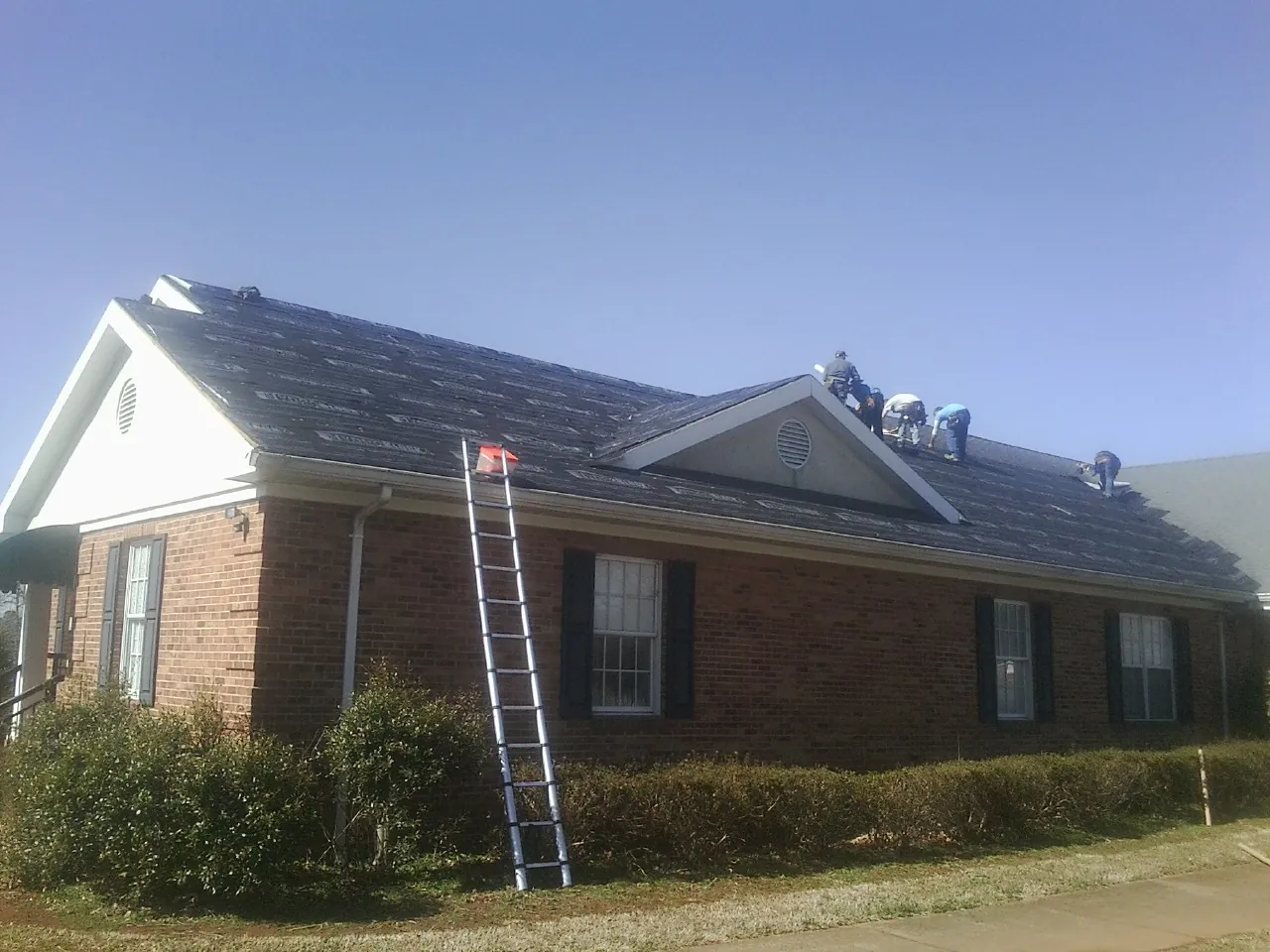 Crew tearing shingles off a brick ranch roof with a ladder set — Emergency Roof Repair work by our crew in Benge