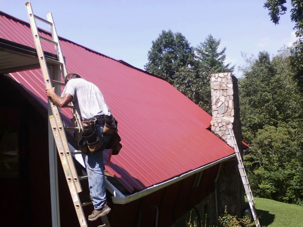 Roofer on a ladder beside a red metal roof and stone chimney — Soffit & Fascia Repair by Roofing Brothers in Benge